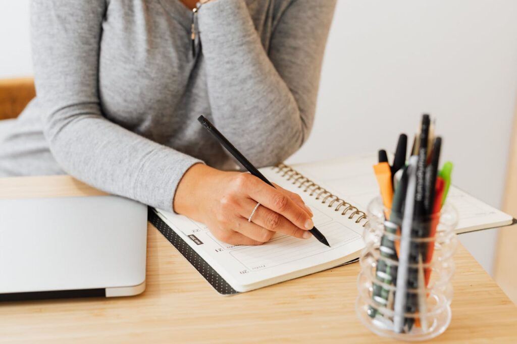 A businesswoman writing in a notebook at a wooden desk with pens and a laptop.